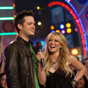 Damien Fahey and Hilary Duff at a vibrant MTV award show. Neon lights and cheering crowd in background.