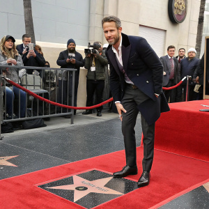 Ryan Reynolds, standing in a black tuxedo, accepts his star on the Hollywood Walk of Fame. Golden star embedded in the pavement, fans and paparazzi in the background.
