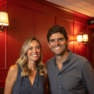 Mark Philippoussis and Amanda Salinas smiling in a cozy, warmly lit room. The background is a rich red. Both are Caucasian.