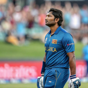 Kumar Sangakkara, Sri Lankan cricket player, in blue team uniform. Serious expression. Cricket field background with spectators in stands.
