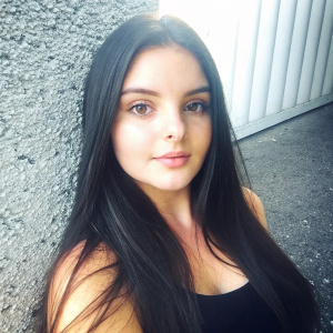 Caucasian actress Ariel Winter in a candid snapshot. Long dark hair, natural makeup. Wearing a black top. Neutral toned wall in the background. Soft lighting