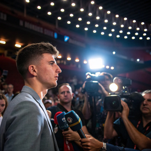 Mason Mount at the press conference, dressed in a casual suit, surrounded by media and fans in a brightly lit auditorium.