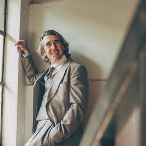 Steve Coogan, British actor with gray hair and a charming smile, in a formal gray suit. Posing against a backdrop of a plain white wall marred by a few stains.