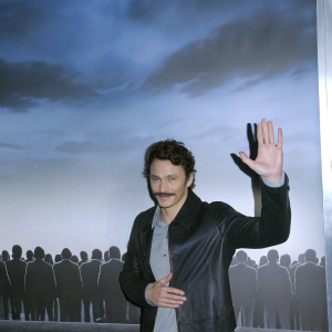 James Franco, known for his luxuriant mustache, posing with a warm and benevolent expression. A sea of stylized silhouettes against a cerulean backdrop. Photo from a memorable event.