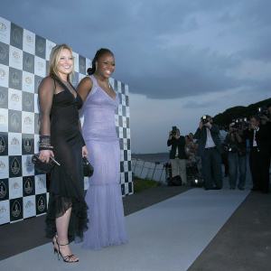 Hilary Duff and Serena Williams at a charity gala. Hilary in a stunning lavender gown with sheer fabric and choker detail, Serena in a sleek black dress. Smiling for photographers against a checkered backdrop with sponsor logos.