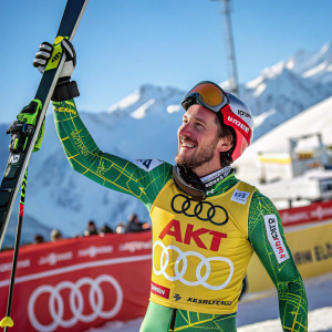 Marcel Hirscher, wearing a green and yellow ski suit, smiling broadly, holding ski poles. Winter sports event backdrop