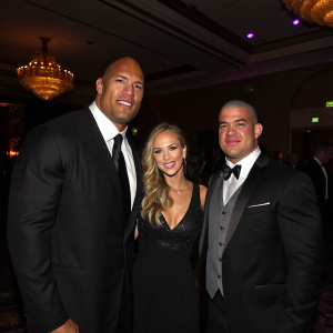 Dwayne Johnson, Amber Nichole Miller, and Tito Ortiz at a gala. Dark suits, formal attire, dimly lit ballroom