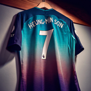 High-definition close-up image of Heung-Min Son's Tottenham Hotspur jersey. Teal-green and navy-blue color scheme. Number 7 prominently displayed on the front. Jersey hung on a hanger in front of a light-colored wall