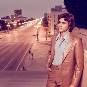 James Franco in a stylish brown leather jacket, light blue shirt, standing alone on a bustling city street at night.