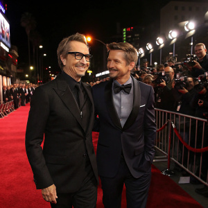 Gary Oldman and Jeremy Renner at a film premiere in downtown Los Angeles. They are holding hands and posing for the cameras. The red carpet is lined with flashing lights and paparazzi.
