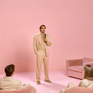 Roger Federer, in a formal suit, giving an interview. Bright lights, studio setting, audience present.