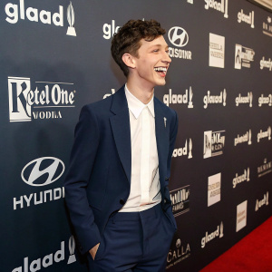 Troye Sivan at the GLAAD Media Awards. Smiling widely on red carpet. Wearing navy blue blazer and white dress shirt. Dark backdrop with sponsors' logos including Coca-Cola, Delta, Barilla, and GLAAD