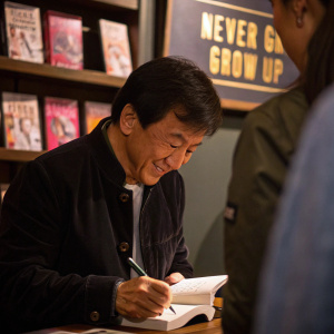 Jackie Chan signing autographs at the book launch event for 'Never Grow Up'. Dark ambient lighting, close-up shot.