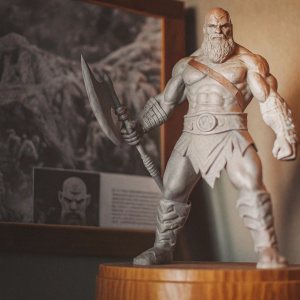 An intricately detailed bronze statue of Kratos from God of War. The statue stands in a dynamic, battle-ready pose with a fierce, determined expression. The lifelike musculature and detailed textures are set on a wooden base adorned with an informative plaque.