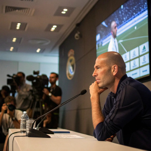 Zinedine Zidane in a contemplative mood at a press conference. Large screen showing match highlights in the background.