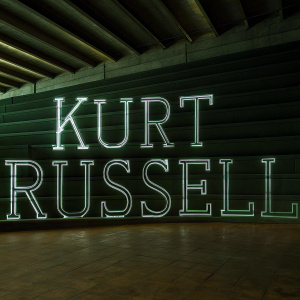 Large, sleek font announcing Kurt Russell's participation in the upcoming blockbuster. Modern design, monochrome theme, promotional material.