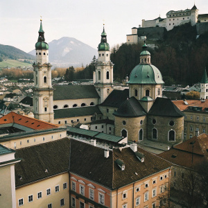 High-altitude aerial view of Salzburg's historic district. Dominant structures include the towering spires of the Dom Cathedral and the Franciscan Church, set against a dramatic mountainous backdrop.