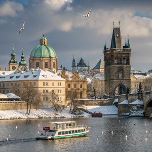 Winter scene of Prague, Czech Republic. Ornate architecture of Gothic and Baroque styles visible. Charles Bridge and Old Town area seen from the Vltava River. Snow covers some buildings. A boat and birds on the calm river. Partly cloudy sky