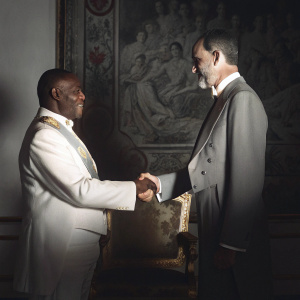 Umaro Sissoco Embalo and King Felipe VI, in elegant suits, greet each other. Golden throne room, ornate tapestries with ancient figures.
