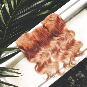 Close-up photograph of blonde hair extensions. Wavy body wave style, light blonde color. White background highlighting natural voluminous appearance.