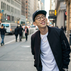 Jay Park, Asian male, casual pose with friendly demeanor. Wearing a black jacket over a white t-shirt, glasses and a baseball cap. The background is a bustling urban street.