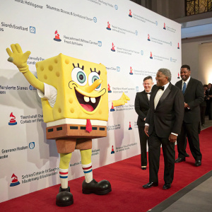 SpongeBob SquarePants character, in mascot costume, standing alongside formally dressed guests at a Smithsonian National Postal Museum event. White backdrop with event logos.