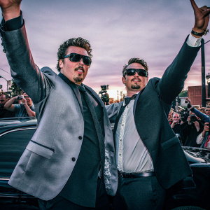 Danny McBride and James Franco at a movie premiere. Wearing sharp suits and dark sunglasses. Crowds cheering in background.
