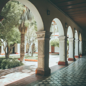 The serene courtyard of Saint Catherine's Monastery in Arequipa, Peru. Filled with lush greenery and bathed in sunlight. Archways and columns with intricate designs. Path paved with brick, vibrant hues of white and brick-red. A blend of colonial charm and ornate details.