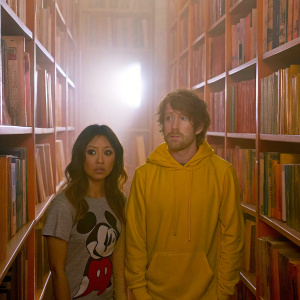 Brenda Song and Seth Green in a cozy bookstore scene. Both have serious expressions. Brenda is in a gray shirt featuring Mickey Mouse, Seth is in a yellow hoodie.