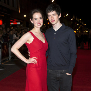 Anna Popplewell and Skandar Keynes posing at a film festival. Anna wears a stunning red dress with no sleeves. Skandar is in a dark long-sleeved shirt. The background features a dimly lit crowd.