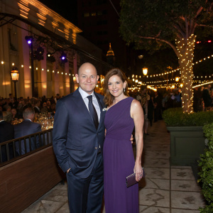 Rob Corddry and Sandra Corddry, at a charity gala. Rob in a sleek navy suit. Sandra in a chic floor-length purple dress. Decorated outdoor venue, spotlights and crowd