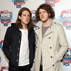 Jamie Cook and Nick O'Malley from Arctic Monkeys at an awards show. Jamie with long, straight brown hair, dark jacket over white t-shirt, focused expression. Nick has curly brown hair, light-colored, structured trench coat. Backdrop with splattered paint and NME Awards logos.