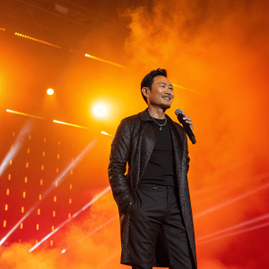 Jacky Cheung Hok-yau at a media event for his concert tour. Bright lights and a fiery orange backdrop behind him.