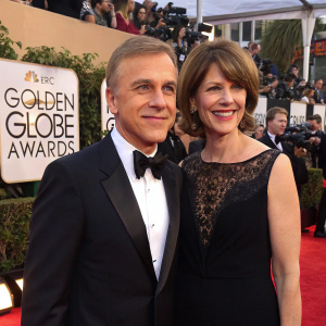 Christoph Waltz and Judith Holste, both in formal attire, at the Golden Globe Awards. Red carpet event with flashing cameras and excited fans in the background.