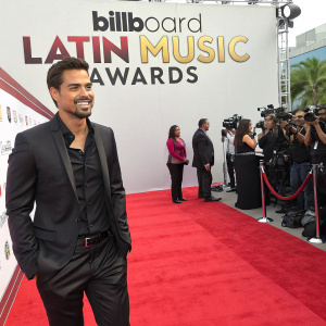 J.R. Ramirez on the vibrant Billboard Latin Music Awards red carpet. White backdrop featuring logos and sponsor names. Wears a sleek black suit and dark shirt.