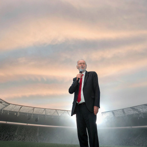 Jeremy Corbyn, in a black suit with red tie, addressing supporters. Crowded stadium, green backdrop