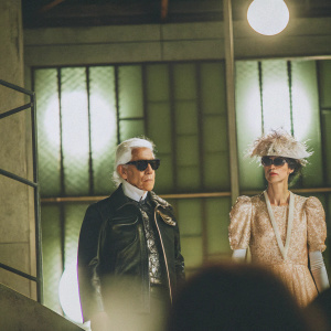 Stella Tennant and Karl Lagerfeld at a fashion gala. The iconic designer wears a black leather jacket and sunglasses, while Tennant is adorned in an exquisite gown with a delicate feathered hat. Their formal poses are set against a modern, industrial backdrop with large lighting fixtures.