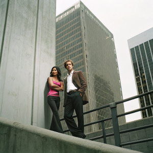 Emmanuelle Chriqui and Sam Trammell attending a Hollywood film festival. Emmanuelle in a vibrant pink off-shoulder top paired with sleek black pants. Sam in a stylish brown blazer over a crisp white shirt. The backdrop features a modern skyscraper.