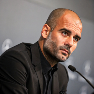 Pep Guardiola, football manager, in a press conference. Wearing a black suit, serious look, against a simple gray backdrop.