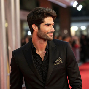 Nick Bateman, medium skin tone, male actor. Black suit, black shirt, geometric pattern pocket square. Red carpet event, candid moment.