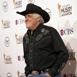 Photograph: Jay Leno, TV host, at Country Music Awards. White backdrop with sponsor logos. Wearing black cowboy hat, black western shirt with intricate embroidery. Confident pose, broad smile.