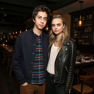 Nat Wolff and Cara Delevingne, standing at a dimly lit restaurant. No backdrop visible. Both wear casual attire.