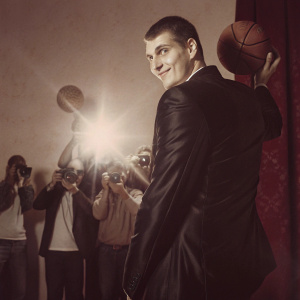 Nikola Jokic, the Denver Nuggets star, in a sleek black suit, posing confidently at the NBA Awards. Surrounded by photographers and fans in a vibrant, star-studded event.