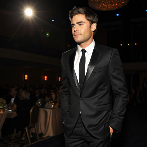 Zac Efron in a sleek black suit with a classic black tie, posing confidently at an award show. The room is dimly lit, with blurred audience in the background.