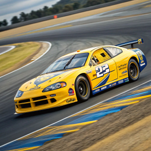 Dodge Intrepid R/T speeding around the Monaco Grand Prix circuit. The car is adorned in vivid yellow with blue accents, sponsor logos and decals cover the sleek, aerodynamic body. A photo capturing the intense race action.