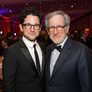 J.J. Abrams and Steven Spielberg, both in sharp suits, at a gala. Abrams, with dark curly hair, and Spielberg, with gray hair and glasses, are in a room filled with people.