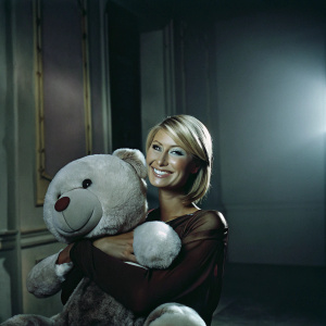 Paris Hilton with a large white teddy bear in her arms, smiling for the camera. Her blonde bob and brown top stand out against a soft, warm background.