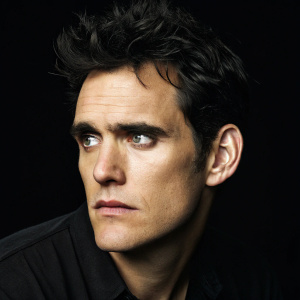 Matt Dillon, intense stare. Close-up portrait, dark hair disheveled. Solid black background.