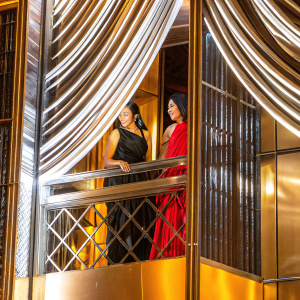 Charli D'Amelio and Avani Gregg at the Met Gala. They are both in formal attire, Charli in a black gown, Avani in a red dress. Luxurious velvet curtain backdrop.