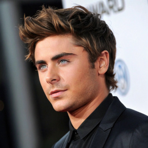 Zac Efron, handsome actor with tousled brown hair, blue eyes, and a chiseled jawline. He's dressed in a sleek black shirt. Shot against a soft, blurred, dark background at an awards ceremony.